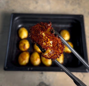 Ugnsform i mörkblått med non-stick, grillad kycklingbit på tång och rostade tillbehör.