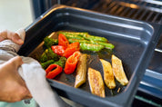 Svart ugnsform med galler, fylld med grillade grönsaker